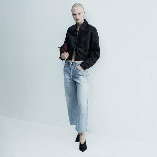 Eine Frau in schwarzer Lederjacke und Jeans steht vor einem Schaufenster 