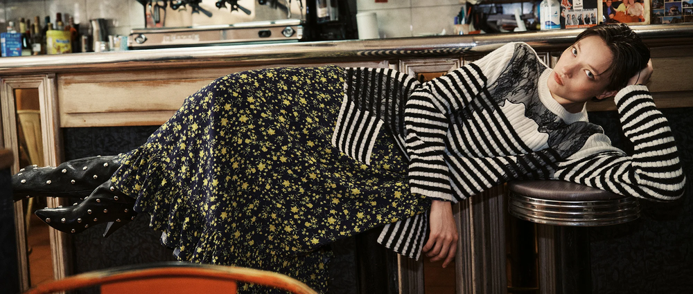 Eine Frau mit welligen, braunen Haaren trägt einen limettengrünen und pfirsichfarbenen Pullover von Essentiel Antwerp