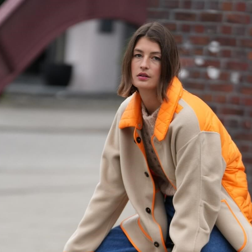 Eine Frau mit einem Mantel mit orangenem Detail sitzt auf einer Bank an der Straße