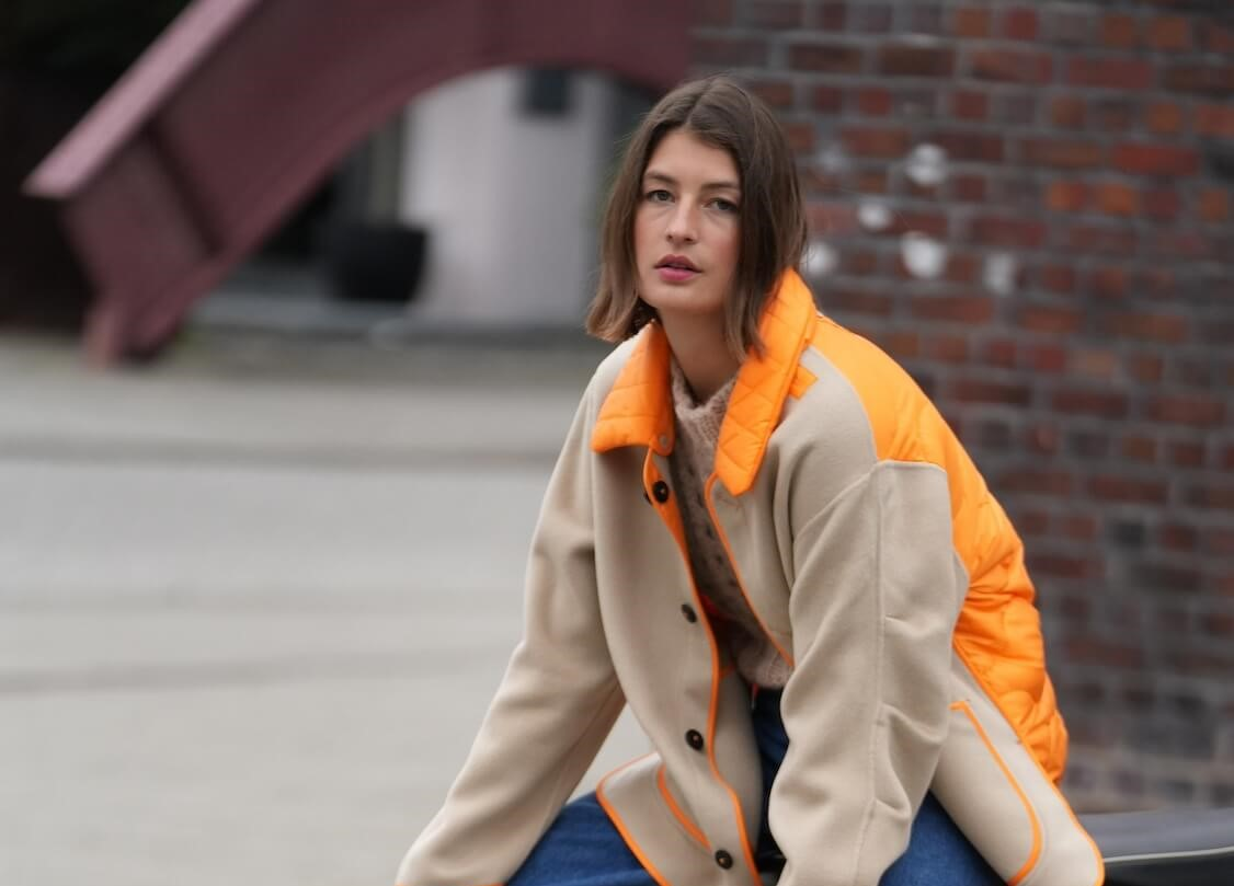 Eine Frau mit einem Mantel mit orangenem Detail sitzt auf einer Bank an der Straße