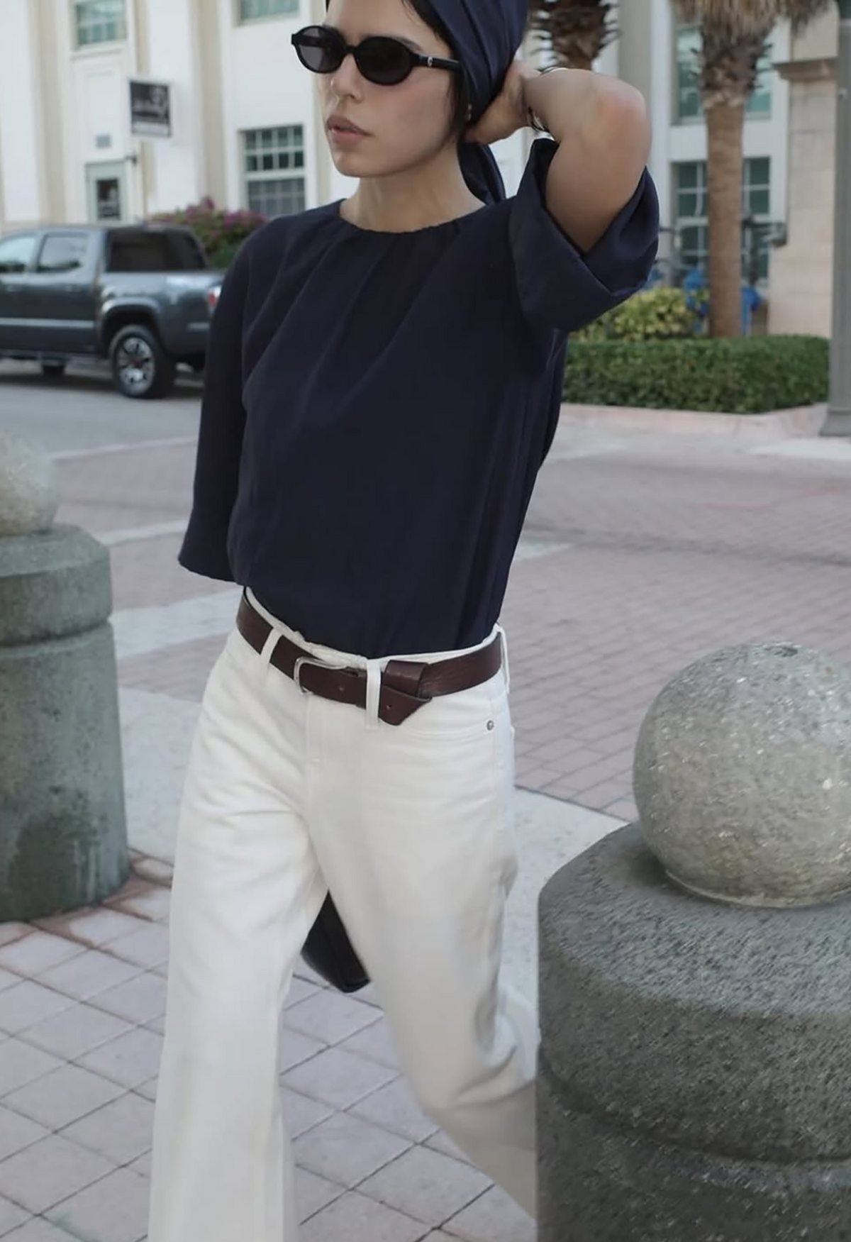 Model trägt ovale schwarze Corlin Sonnenbrille mit dunkelblauem Oversized-Top, weißer Jeans und braunem Ledergürtel im Street-Style-Look – Damen Accessoires bei Marandino