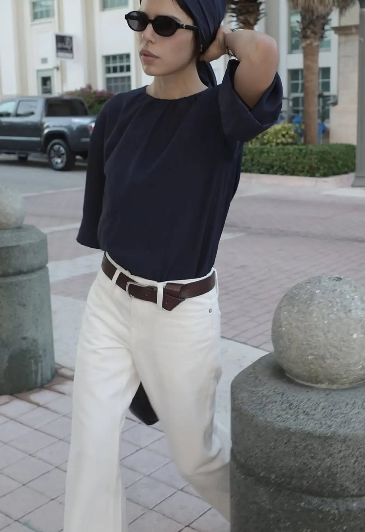 CORLIN Model trägt runde, schwarze Sonnenbrille und ein simples Outfit mit navy Shirt und weißer Hose - sie läuft in der Stadt auf der Straße