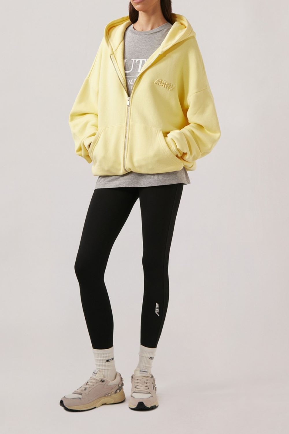 Frau trägt gelbe Kapuzenjacke, graues T-Shirt, schwarze Leggings mit hohem Bund und weiße Sportsocken mit hellen Sportschuhen