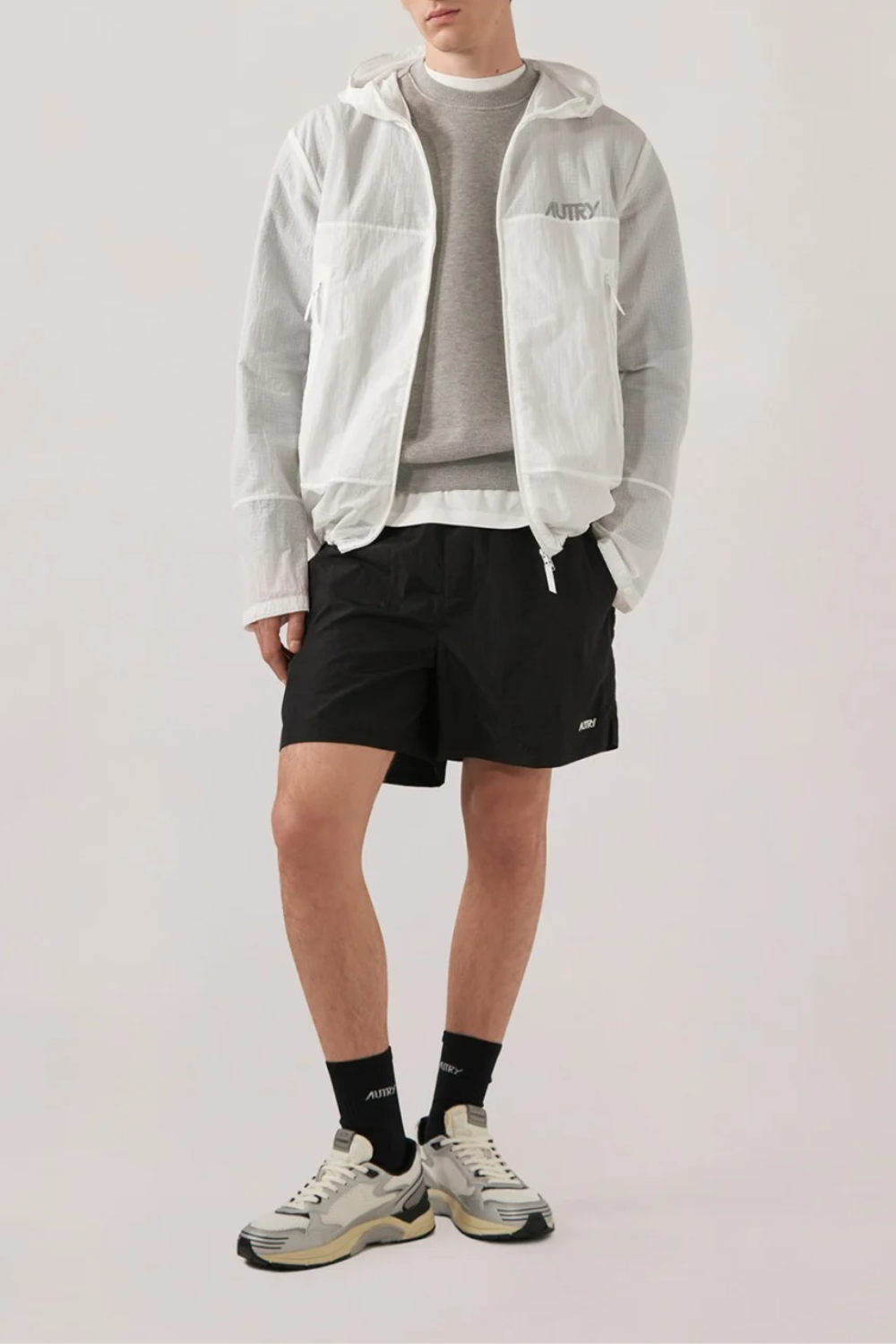 Mann trägt weiße Ripstop-Jacke mit durchgehendem Reißverschluss und Logo-Zipper, graues Sweatshirt, schwarze Shorts, schwarze Socken mit weißem Logo und weiße Sneaker.