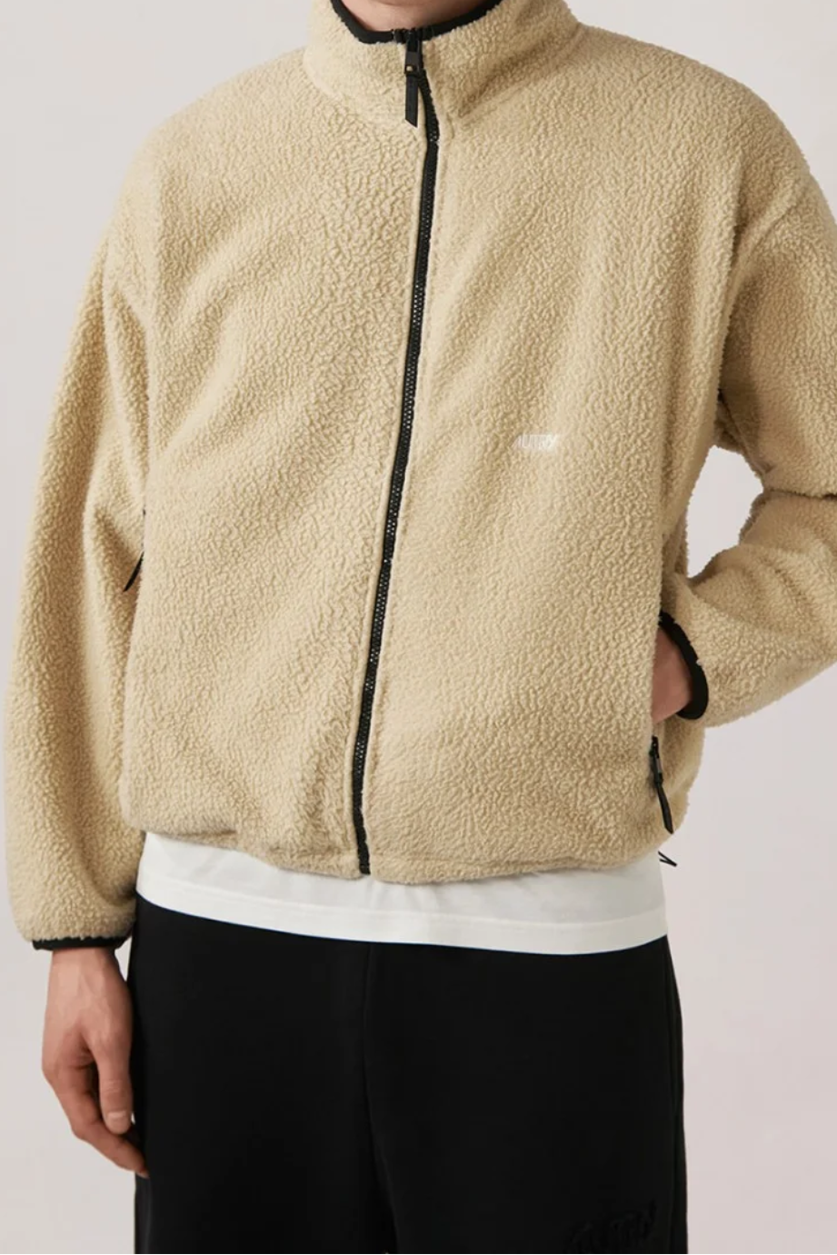 Beige Teddy-Jacke aus 100% Polyester mit durchgehendem schwarzem Reißverschluss, Logo-Stickerei auf der Brust, Kontrastdetails an Ärmeln und Saum, Oversize Fit, getragen von einer Person mit schwarzer Hose und weißem Shirt darunter