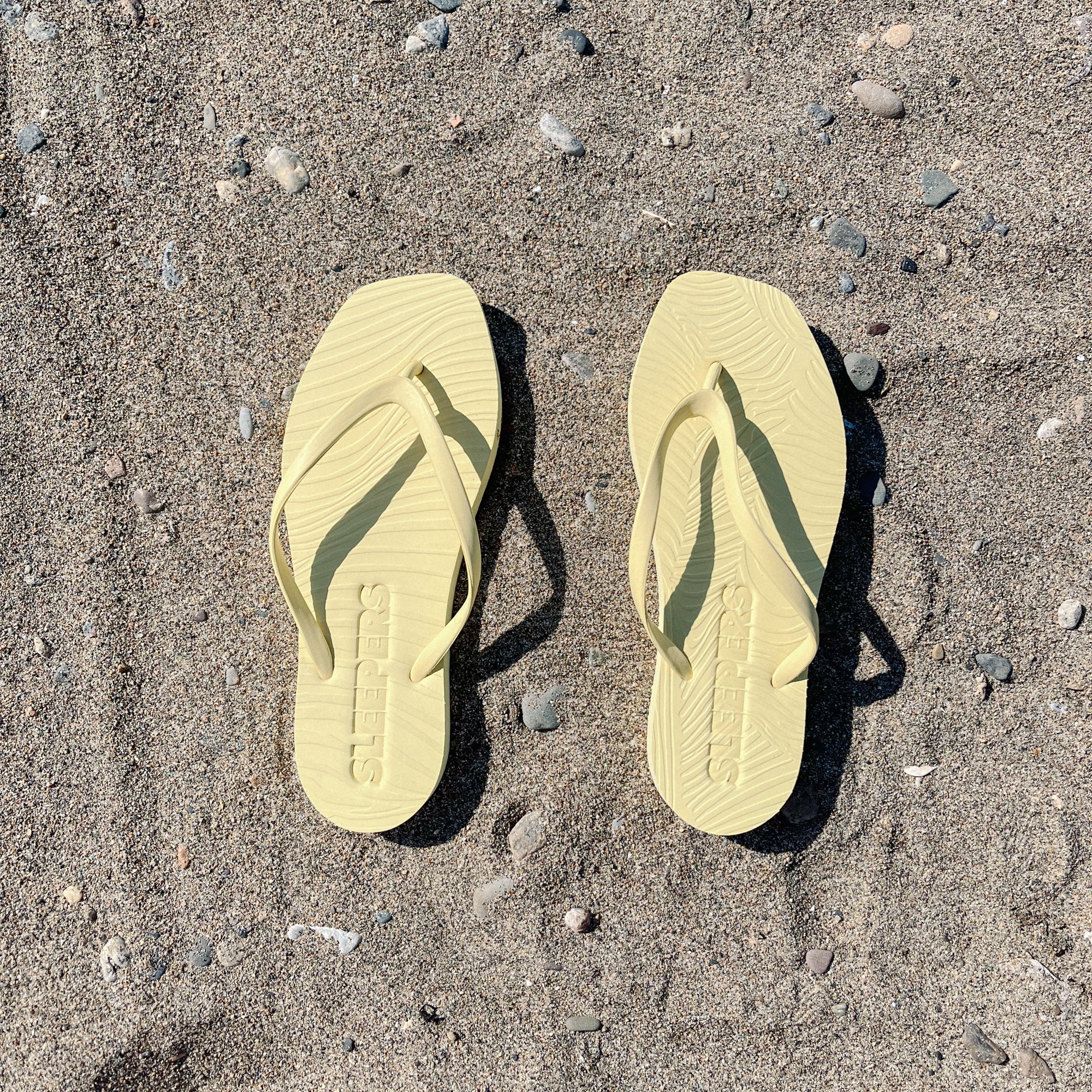 Gelbe Flip-Flops mit gewölbter Sohle und Riemen aus Naturkautschuk liegen nebeneinander auf sandigem Untergrund