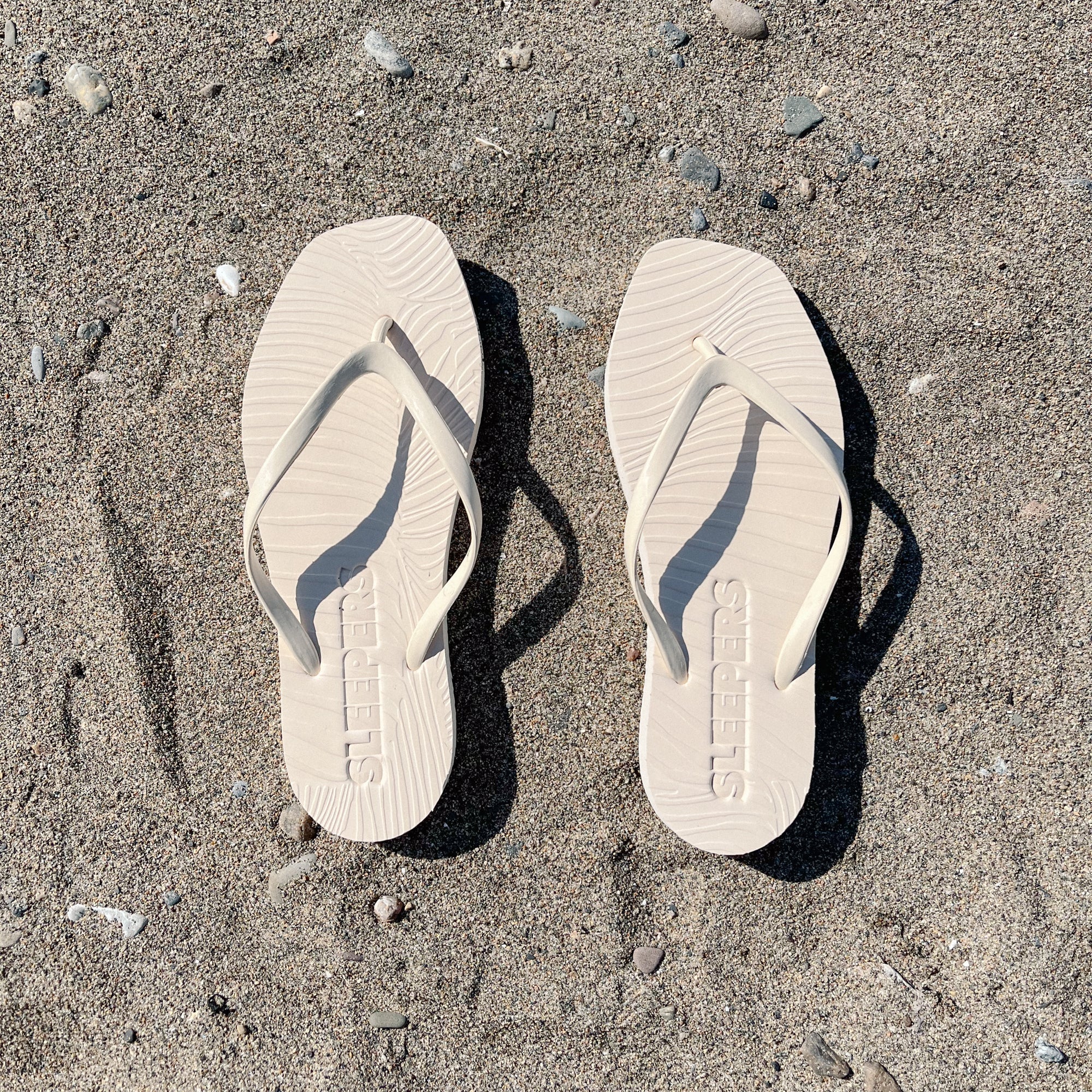 Beige Flip-Flops mit klassischer Silhouette und gewölbter Plateausohle aus Naturkautschuk liegen nebeneinander auf Sand mit kleinen Steinen