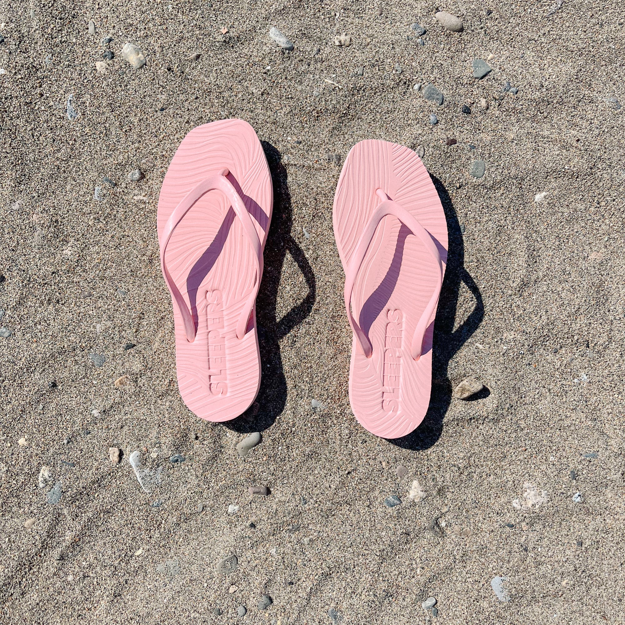 Zwei rosa Flip-Flops mit gewölbter Sohle aus Naturkautschuk liegen nebeneinander auf sandigem Untergrund