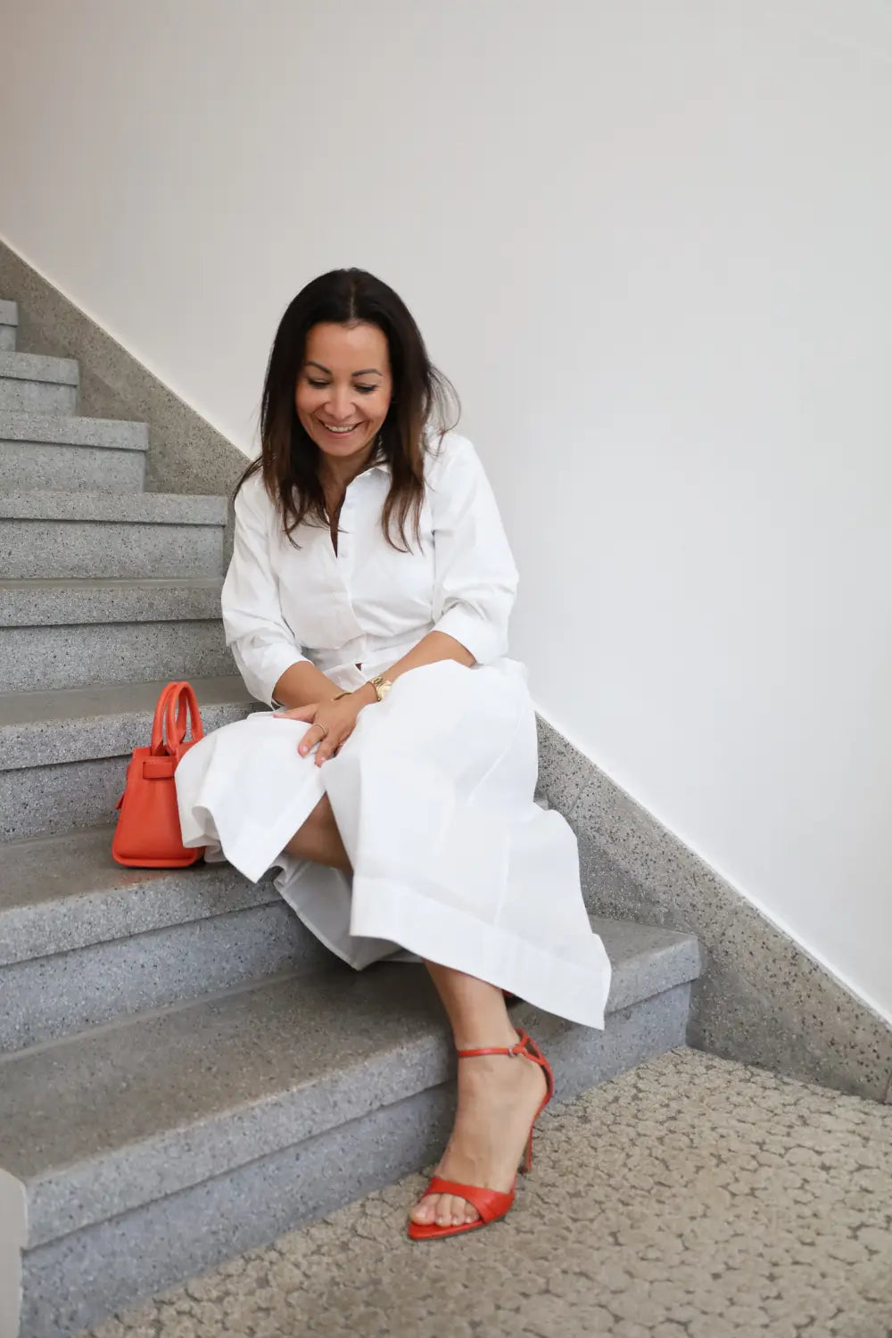 Marandino Inhaberin sitzt auf Treppe in weißem Hemdblusenkleid midi, kombiniert mit roten Sandaletten und roter Mini-Handtasche – Damenmode Sommer-Outfit bei Marandino Nagold