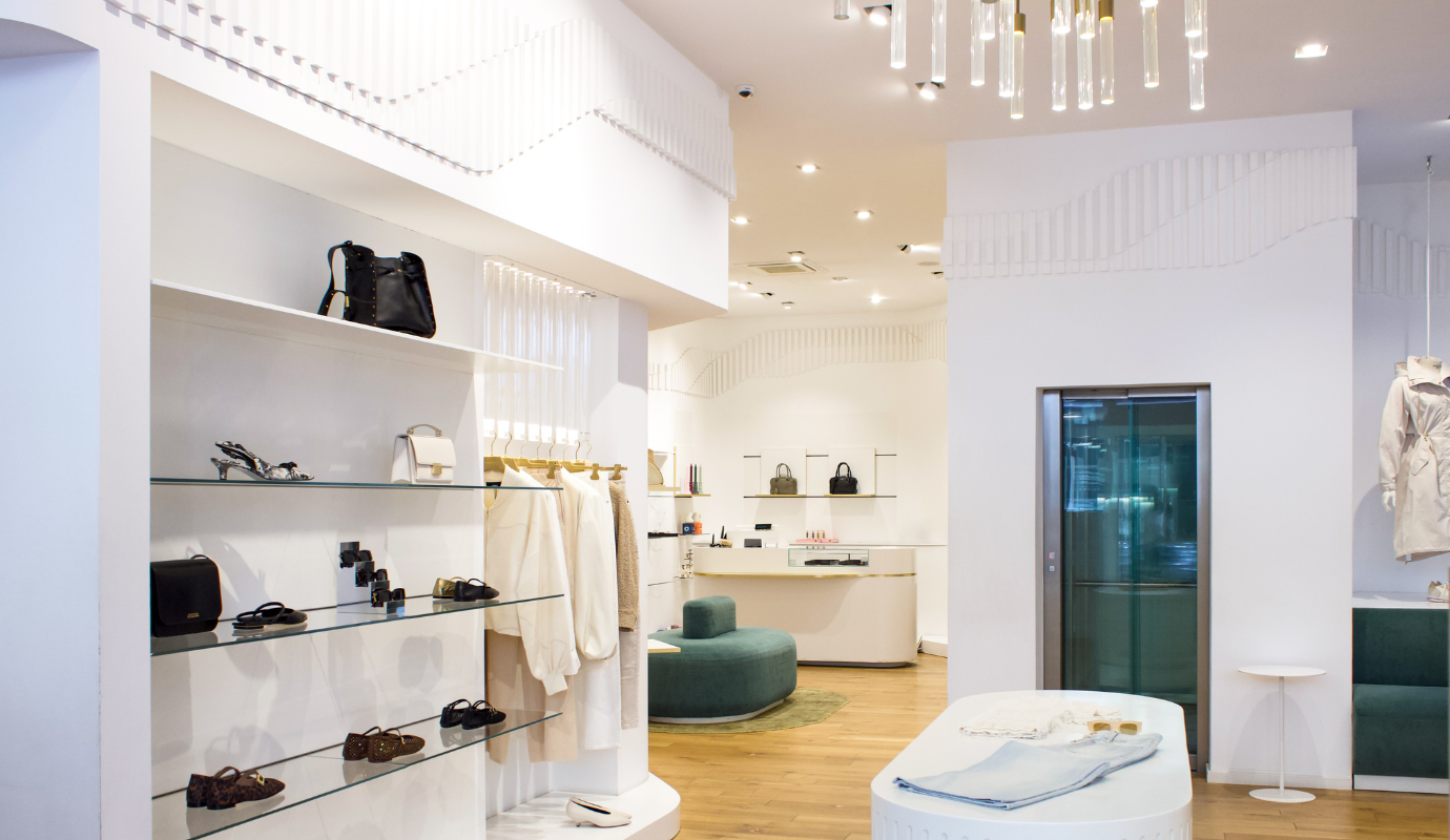 Helles modernes Ladeninnere von Marandino  mit Glasregalen für Schuhe und Taschen, Kleiderstange mit cremefarbener Mode und elegantem Kronleuchter – Modegeschäft